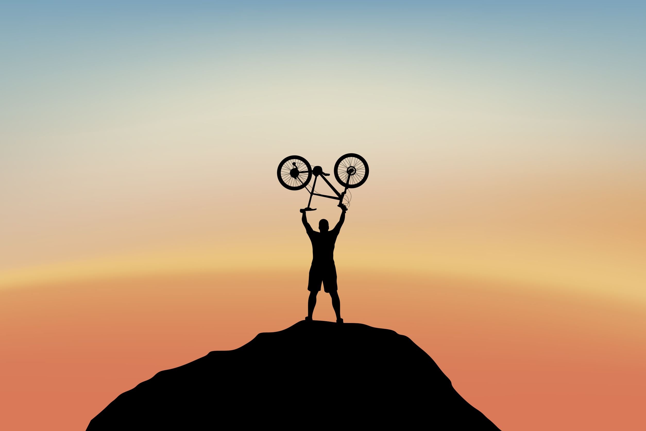 Egy férfi nyerő gesztust tesz, miközben egy mountain bike-ot tart a feje fölött egy domb tetején