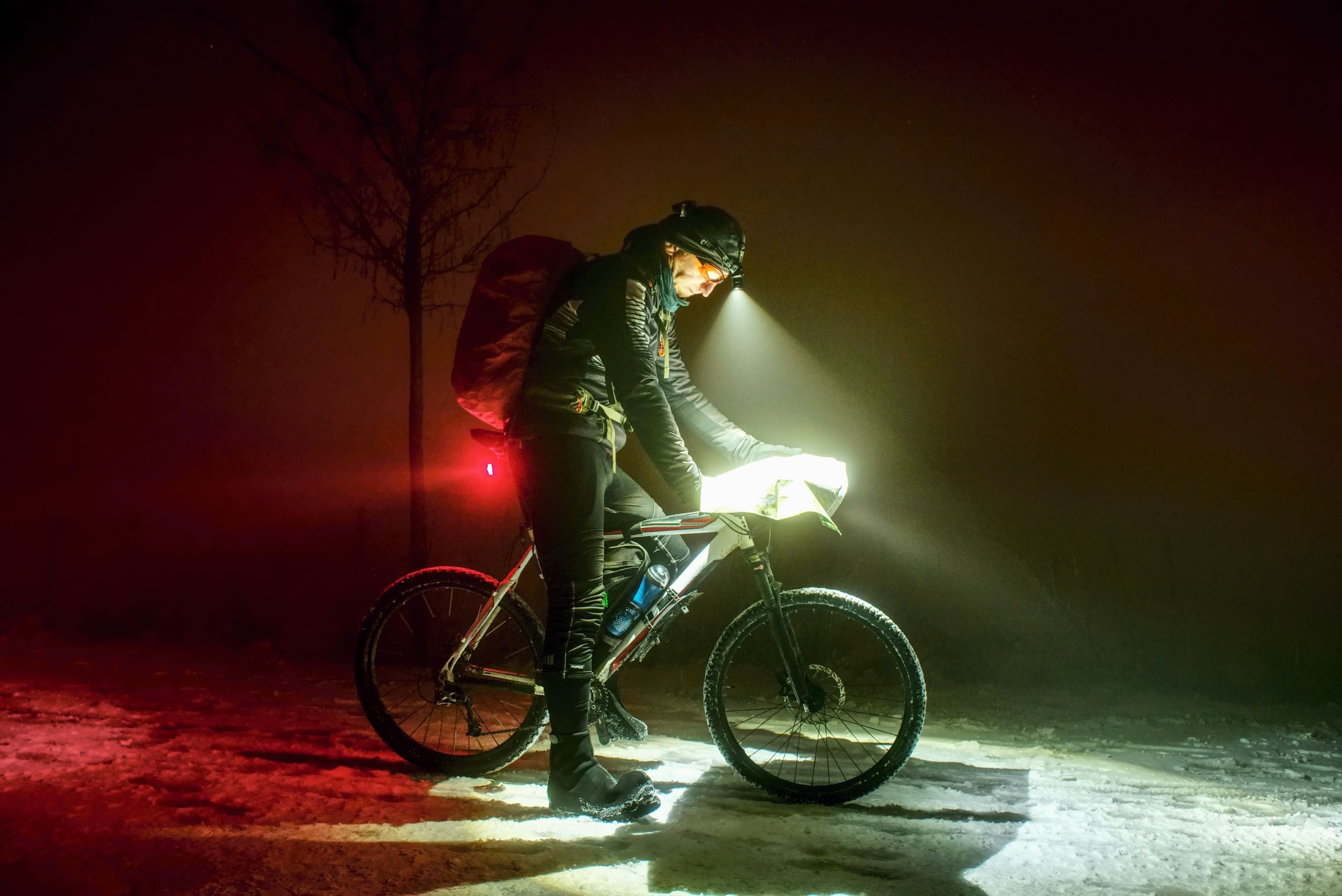 Muž na kole v noci svítí předním bílým světlem, zadním červeným světlem a čelovkou.