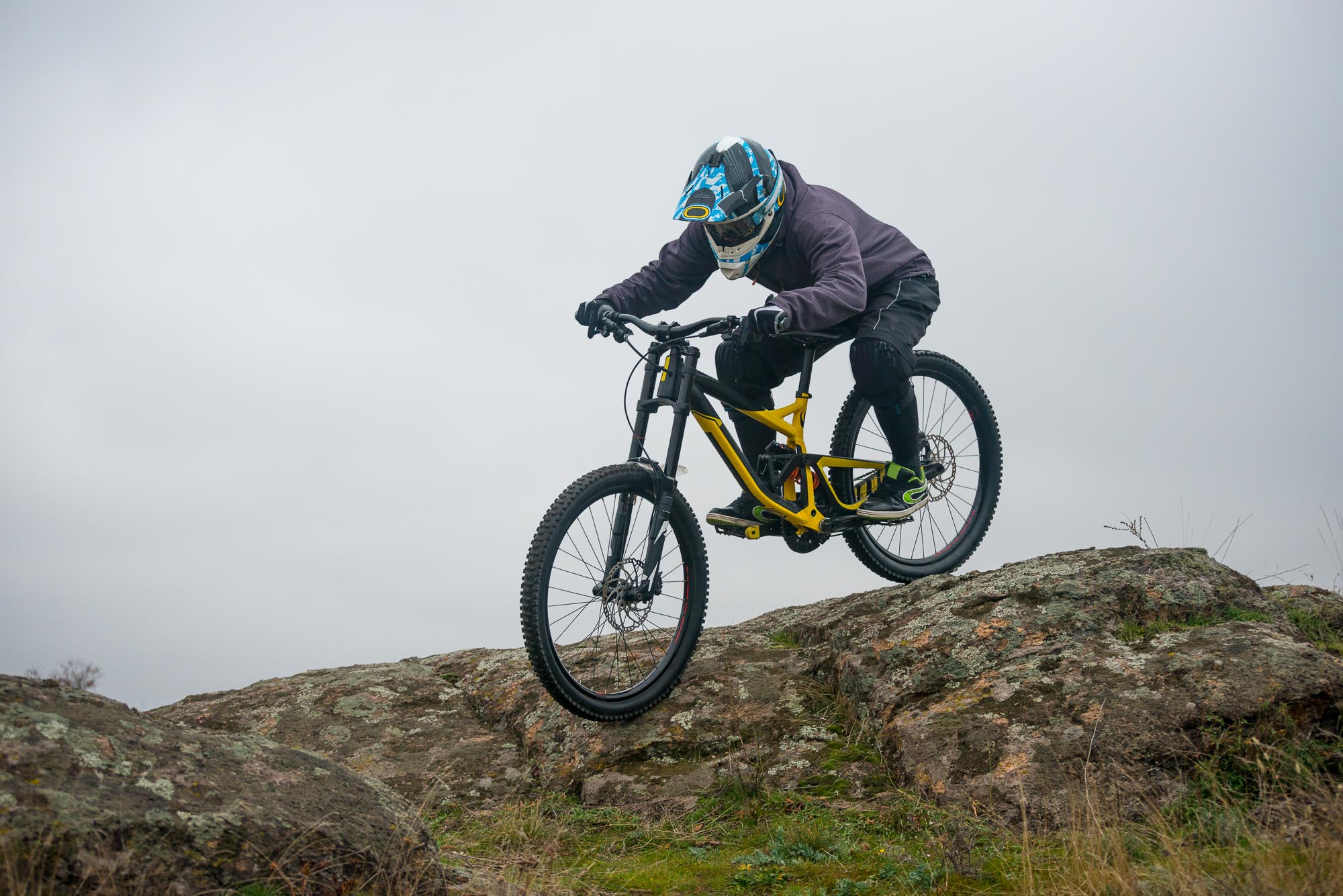 Egy hegyi kerékpáros lefelé halad, mountain bike rövidnadrágot, betétet, lábmelegítőt és kabátot visel.