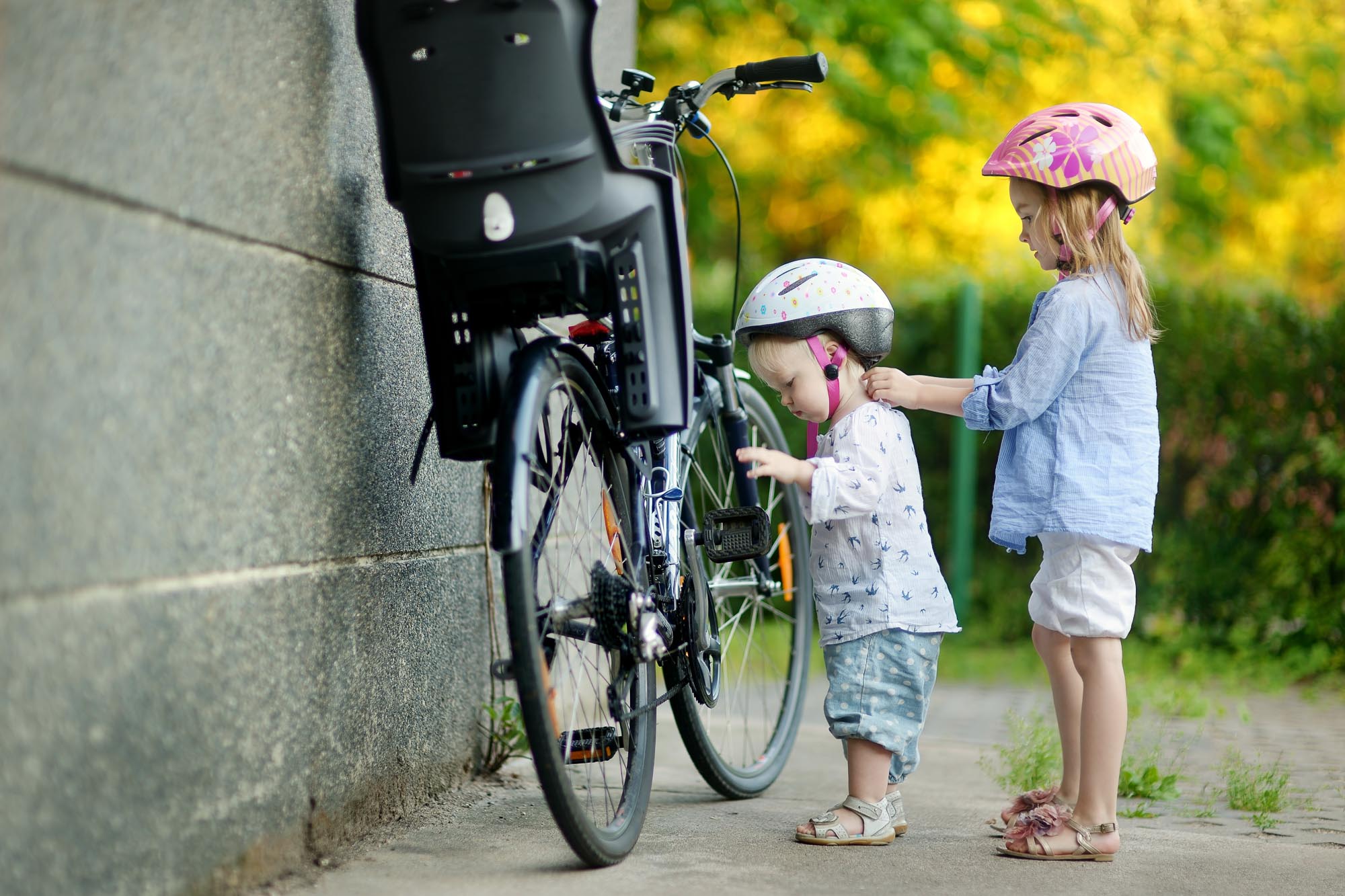 Little girl and taller girl stand by a bicycle with a bike seat.