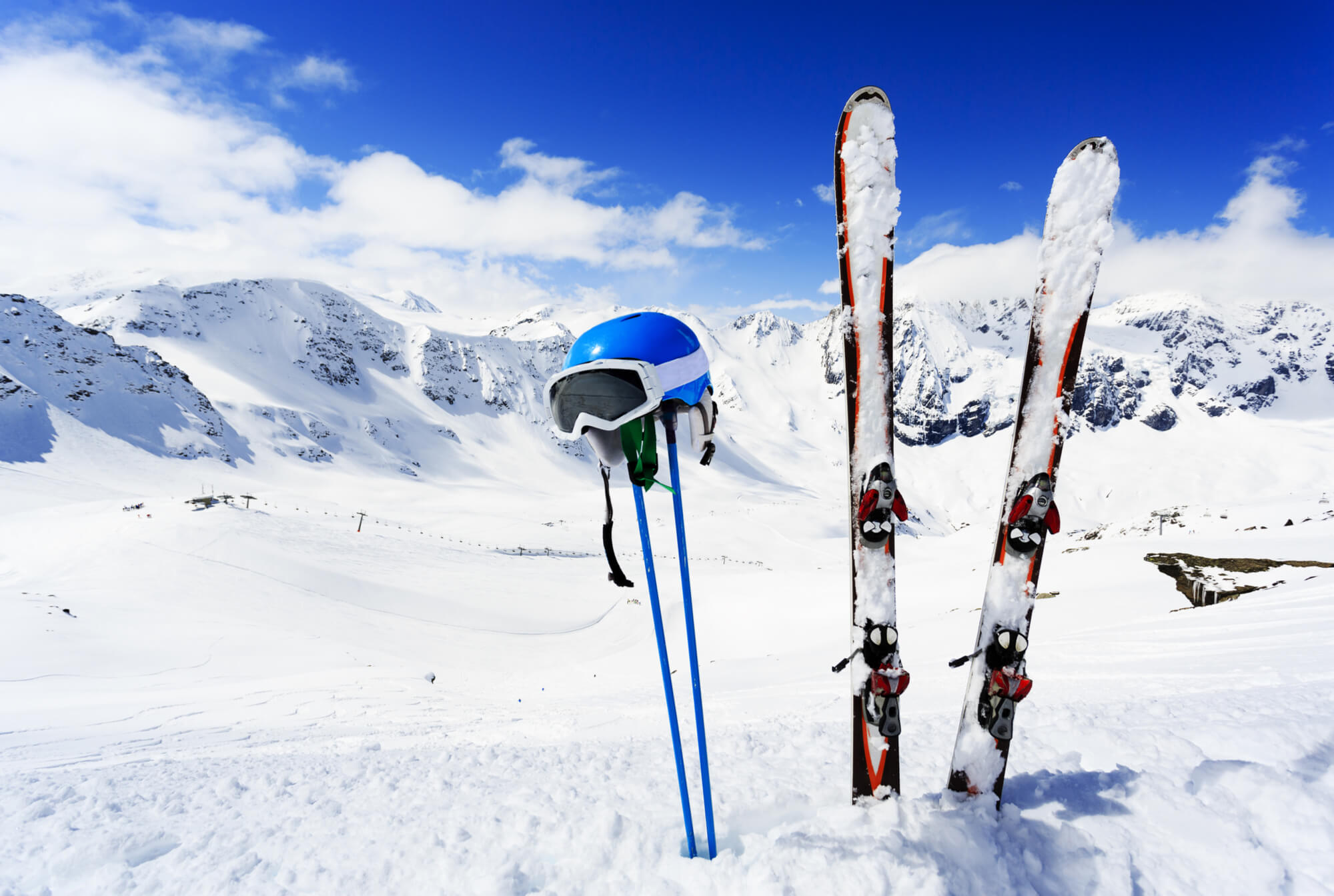 Skis with poles planted in snow and a hanging helmet with goggles with mountains in the background
