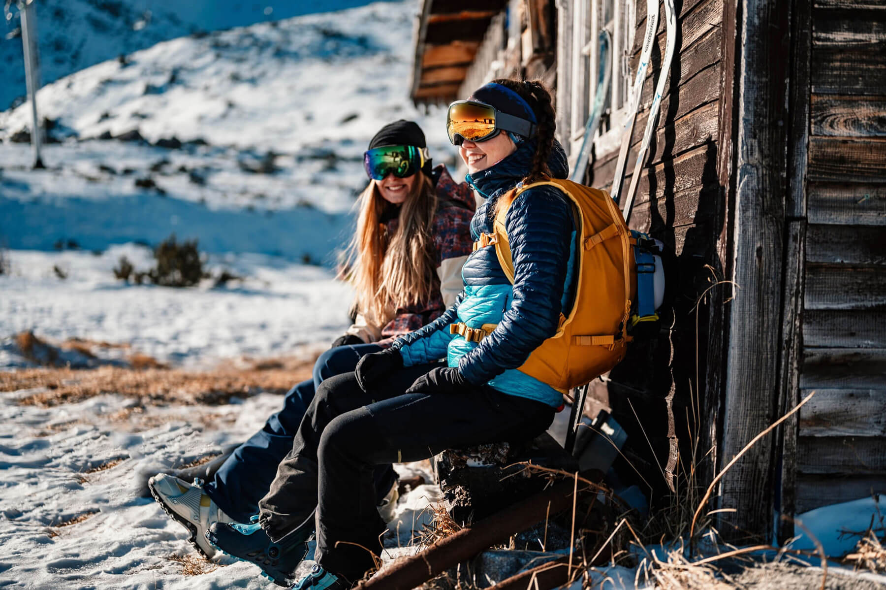 Two ski tourers sit on a bench with skis in the background
