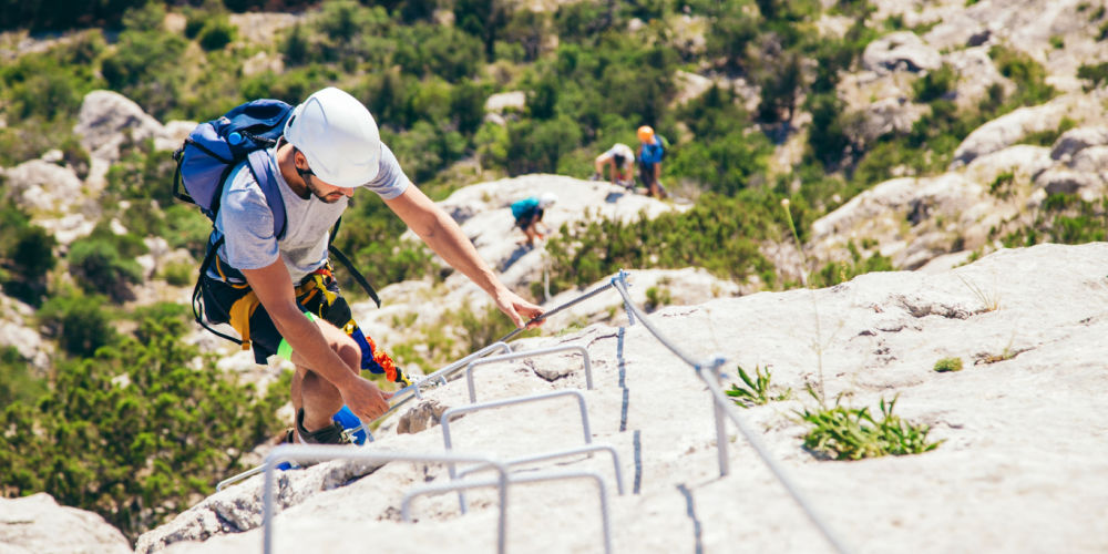 A man with via ferrata equipment is climbing via ferrata, with a group of other people in the background.